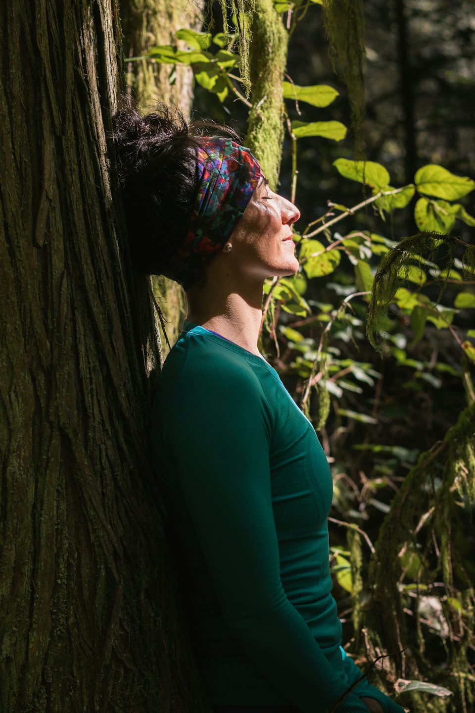 Woman with eyes closed leans against a moss-covered tree in a forest.