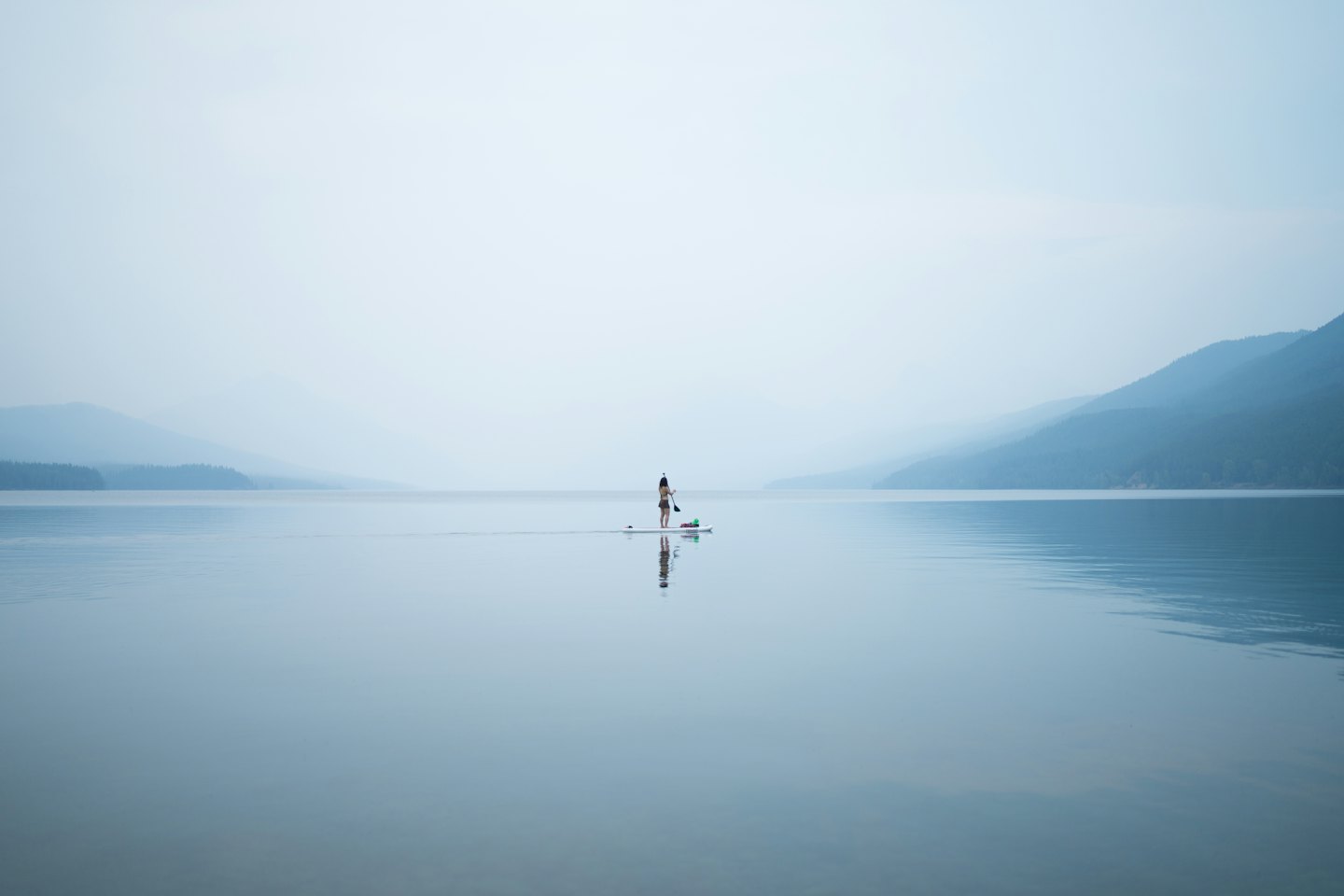 Person paddleboarding across a reflective lake towards distant misty mountains.